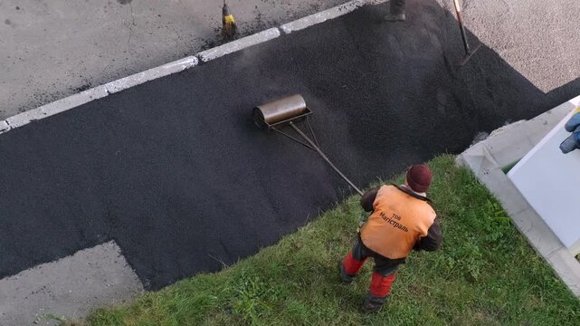 Ukraine, Kiev. June 15 2026. downtown. High angle view of road construction workers in protective vests leveling and compacting fresh hot asphalt on a pathway using a hand roller and rake.