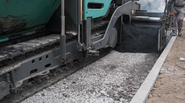Close up of an industrial asphalt paving machine placing a fresh layer of hot black tarmac over a crushed stone foundation on a road construction site.