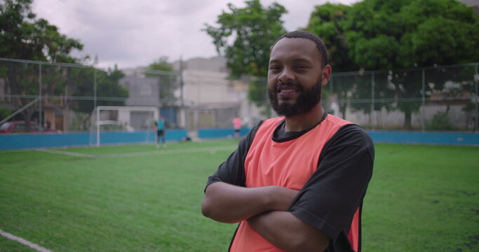 African American Latin man smiling confidently with folded arms on soccer field, teammates playing behind