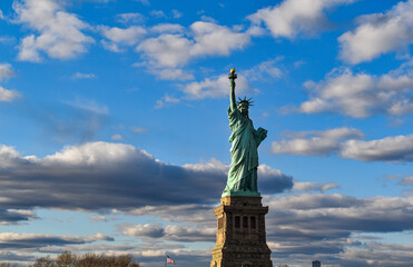 The Statue of Liberty illuminated by warm afternoon light beneath dramatic clouds and blue skies in New York Harbor. A premium commercial travel photograph with generous copy space, ideal for tourism, © Gerard