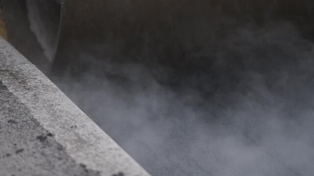 Extreme close-up of a heavy road roller steel drum compacting steaming hot asphalt pavement right next to a concrete curb during sidewalk construction.