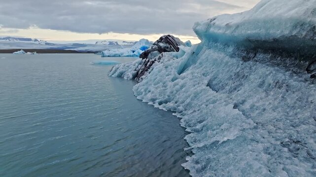 Cinematic aerial flight above floating blue icebergs in J&ouml;kuls&aacute;rl&oacute;n Glacier Lagoon, Iceland, revealing the breathtaking beauty of Vatnaj&ouml;kull glacier and the Arctic landscape at sunset.