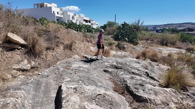 Naxxar, Malta 06.07.2026 - - Salina cart ruts: Two children walk along ancient rock-cut ruts on limestone terrain near residential buildings in Naxxar, Malta. The mysterious grooves, believed to date 