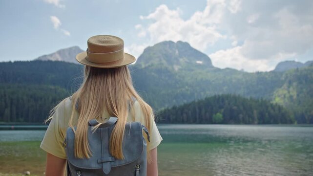 Young woman with backpack admiring Black Lake in Durmitor National Park, Montenegro. Scenic view of the famous glacial lake surrounded by mountains and conifer forest on a summer day