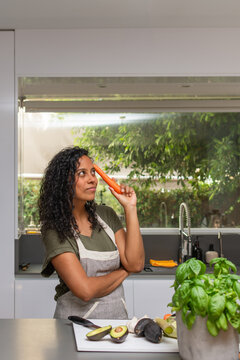 Thoughtful Hispanic woman holding carrot with copy space in modern kitchen