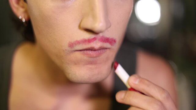 Non-binary person applying red lipstick to draw a mustache and a beard as a form of self-expression
