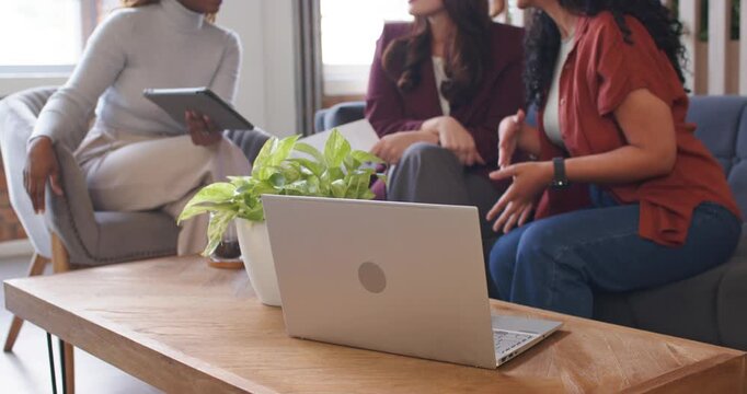 Diverse female team in lounge talking while left woman tapping tablet center holding doc for review
