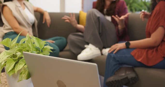 Diverse female friends sitting on couch, gesturing and chatting after camera pullback from laptop