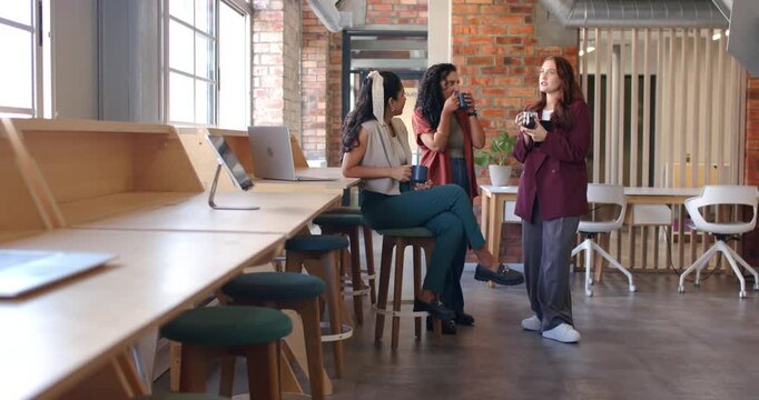 Diverse female coworkers one wearing blazer sipping mugs and chatting at bar counter during break