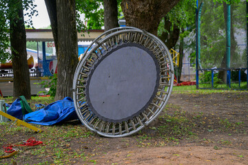 Fototapeta premium Broken trampoline lies on grassy ground. Trees stand nearby in a park setting. Blue tarp and red rope rest beside it. Background shows playground equipment. Metal frame shows signs of wear and tear