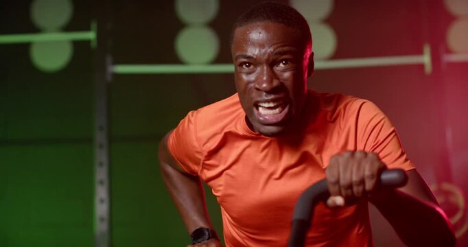 Adult African man gripping cardio bike handles, pulling for interval training in gym in orange tee