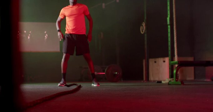 African-American man in orange shirt letting battle rope standing hands-on hips at gym prepping set