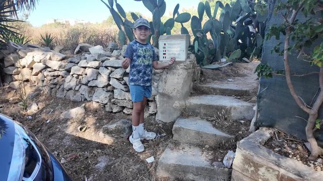 Naxxar, Salina, Malta 06.07.2026 - a young boy wearing a Malta cap points to a multilingual sign reading Footpath through private land, standing beside a stone wall and cactus patch on the path leadin
