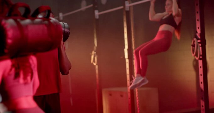 Diverse partners, man carrying sandbag walking forward, rig athlete doing leg raises in circuit gym