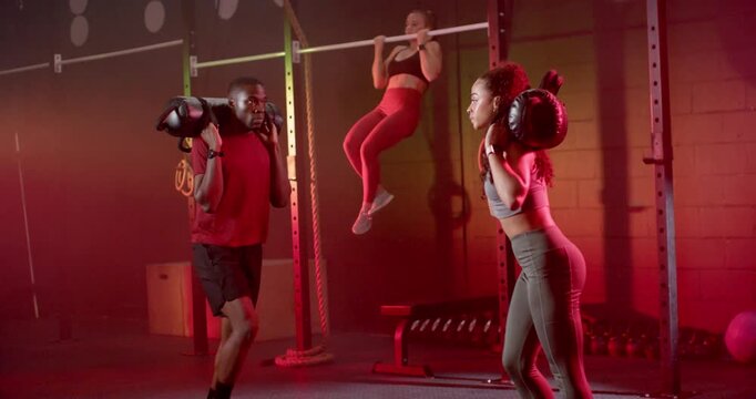 Training partners starting, doing sandbag lunges in gym in sportswear while teammate doing pull-ups