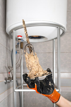 Technician Holding Removed Water Heater Heating Element Covered With Heavy Limescale In Front Of Water Heater. Hard Water Mineral Deposits, Appliance Repair And Replacement Concept.