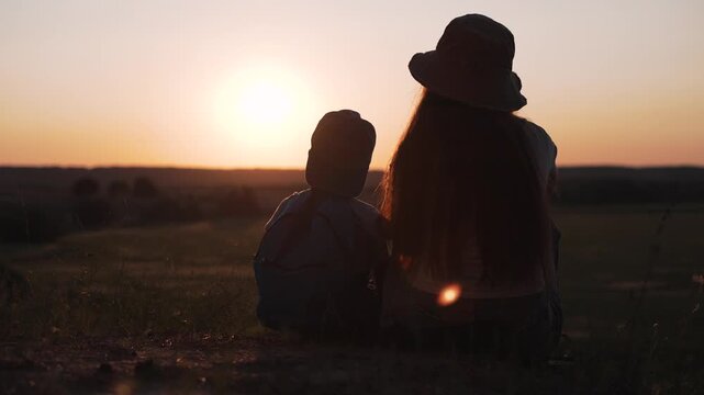 Mother and child sit watching sunrise in countryside field. Woman with boy enjoys golden hour outdoors. Family watches sunrise together. Mother and child bond at dawn. Boy sits with mother in nature.