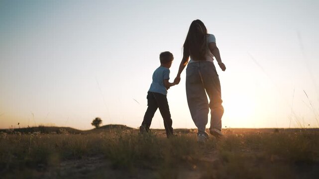 Mother and child walk holding hands with arm raised at sunset. Boy walks with mother in field outdoors. Family silhouette at golden sunset. Child points while walking with mother in field.