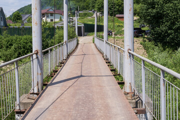 Obraz premium Metal pedestrian suspension bridge in a mountain village with lush green hills. Scenic countryside infrastructure for travel, tourism and transportation concepts.