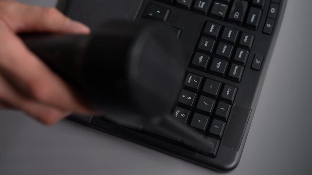 Close-up top view of person using air blower to clean dust from black ergonomic computer keyboard. Focus on numeric keypad. Concept of office hygiene, tech maintenance and workplace cleanliness.