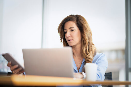 Blond haired mature businesswoman working on her laptop and using a smartphone at office desk