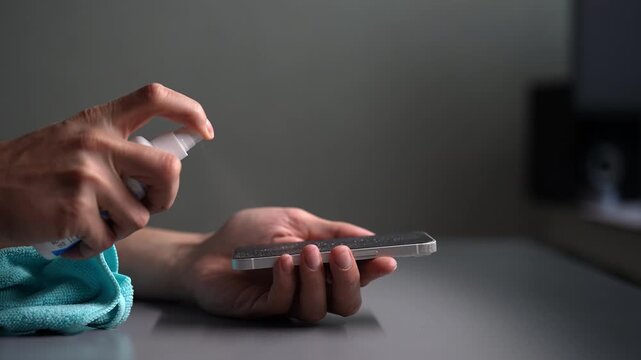 Close-up of hands spraying disinfectant on mobile phone screen and wiping it with microfiber cloth. Concept of device hygiene, sanitation, preventing germ spread. Cleaning electronics.