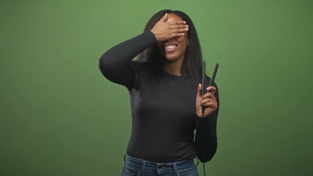Woman holding hair straightener and covers eyes with hand in studio while smiling and wearing black long sleeve top; playful shyness.