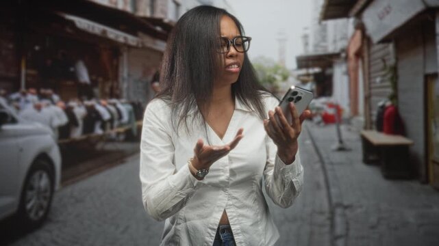 Young african american woman holds smartphone, frowns and gestures with hand while checking the screen on a crowded street market; frustration technical issue.