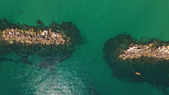 Stunning aerial zenithal view showing a kayaker paddling on a yellow kayak through a passage between two stone breakwaters in beautiful clear turquoise water on a sunny summer day