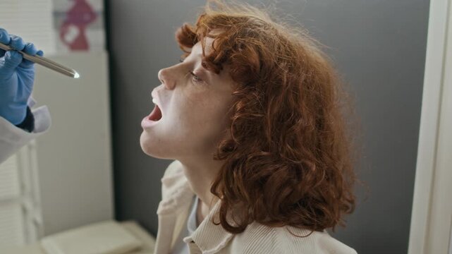 Young Caucasian female patient opening mouth during throat examination with medical penlight in fertility clinic. Ideal for healthcare services, ENT clinic promos, and medical checkup advertisements