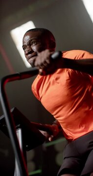 Vertical video: African man pedaling on air bike in gym, gripping handles and pushing to max effort