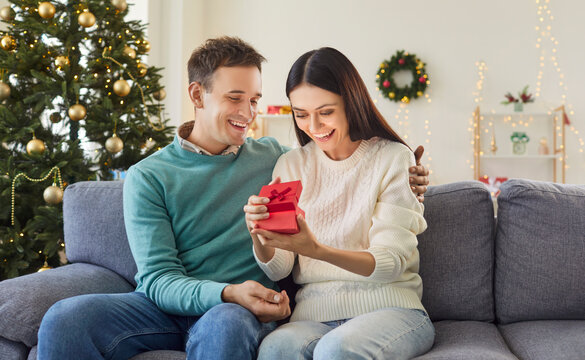 Wife opening a gift box from husband while sitting on the couch during a New Year or Christmas celebration at home. The moment is filled with love, surprise, and festive family joy.