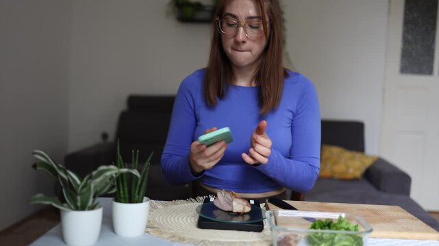 Woman tracking calories weighs cooked chicken on kitchen scale while checking nutrition app on smartphone Preparing a balanced healthy meal with careful portion control daily