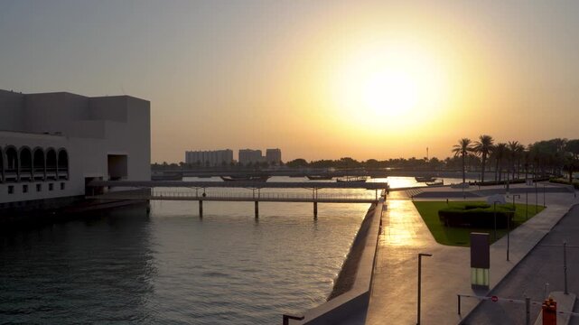 A scenic aerial view of the Doha waterfront at sunset, featuring a pedestrian bridge connecting to a modern building, palm-lined walkways, and traditional dhow boats silhouetted against the glowing.