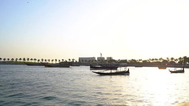 A peaceful view of traditional wooden dhow boats moored in the calm waters of Doha Bay during sunset, with a scenic palm-lined coastline in the background, capturing the quiet essence of Qatar.
