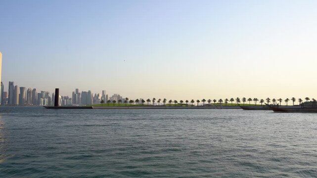 A scenic panoramic view across the bay showing the modern Doha skyline in the distance, set behind a serene waterfront area featuring a continuous row of palm trees and traditional dhow boats.