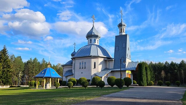 Orthodox church building with silver domes and crosses next to a blue holy water well chapel in a green park with pine forest under blue sky, eastern christian temple architecture
