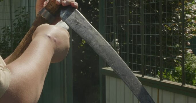 Farmer's hands sharpening a traditional scythe blade with a whetstone, preparing the agricultural hand tool for mowing grass or harvesting crops in the backyard on a sunny summer day.