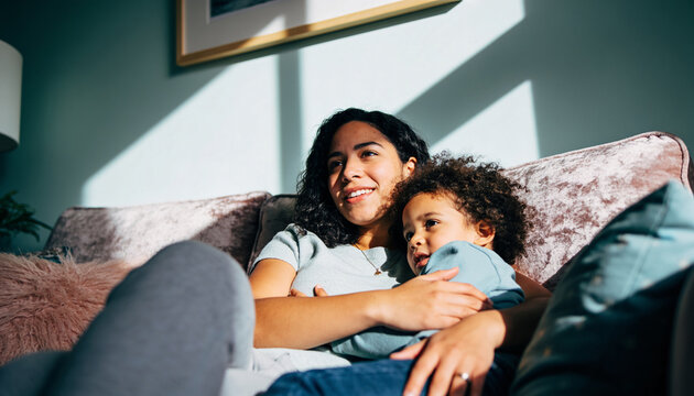 Mother and toddler child cuddling on sofa in sunlit living room with geometric shadows on teal wall indoor