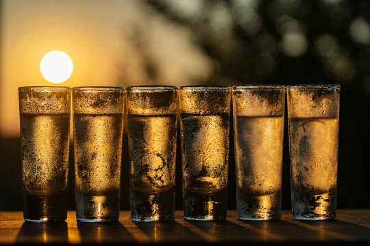 Row of cold shot glasses of vodka or other alcoholic beverages at sunset, closeup. Six chilled alcohol shot glasses filled with clear liquid and covered in condensation drops on golden sunset sky
