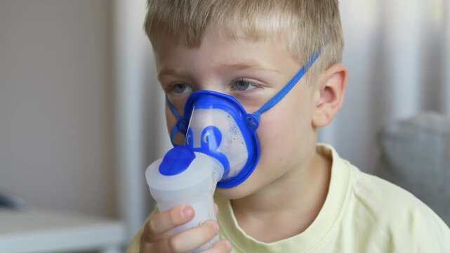 Boy holds nebulizer device himself while breathing through mask looking down in independent home treatment. Child self care and respiratory illness home management concept.
