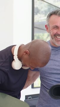 Vertical video: Left man patting shoulder, laughing in room holding green mat, wearing fitted tees