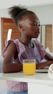 Vertical video: Typing student in purple top studying at kitchen table with laptop and orange juice