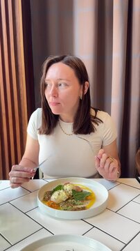 Close-up of a woman eating zucchini fritters with creamy stracciatella cheese, fresh herbs, and tomatoes using a fork and knife at a cafe table