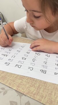 Cute little girl focused on writing English letters on practice sheet with pen at desk for early childhood education and homework stock footage background. High quality 4k footage