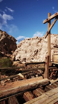 Wooden structure holds a lantern near old mining tools in a rocky landscape. The sun shines, and clouds move in the sky above the site.