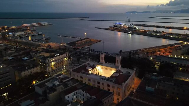 An elegant aerial backward moving drone shot flying diagonally away from the historic Palazzo Civico, revealing a breathtaking wide panoramic view of the sea port, ferry ship, and evening skyline of C