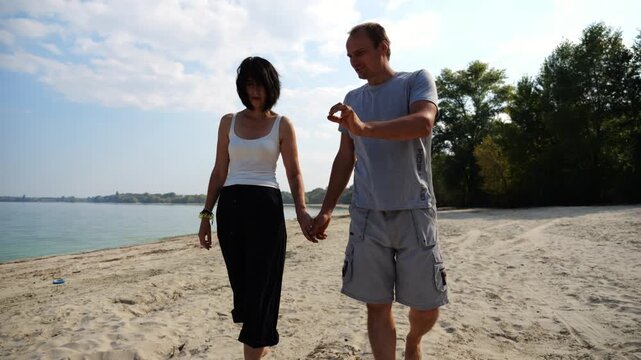 Happy pair going along seashore at sunny day. Romantic couple holding hands of each other and walking at coast. Lovers spending time together at resort. Concept of summer vacation or holiday. Slow mo