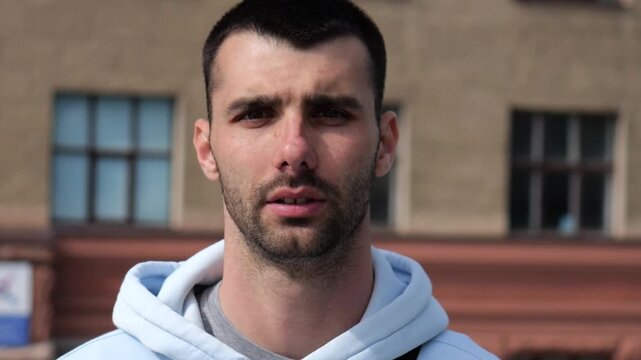Portrait of young confident bearded man in casual clothing with cityscape background. Close up of handsome guy looks into camera standing at city environment at sunny day