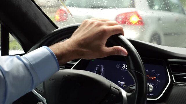 Male hands of businessman holds steering wheel while driving electrical vehicle at rain city. Young man commuting to work at rainy day. Businessperson operating electric car on urban road. Close up